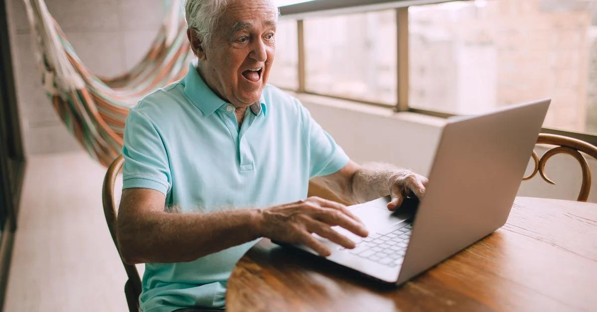 Un homme senior assis à l'intérieur, surpris, utilisant un ordinateur portable sur une table en bois.