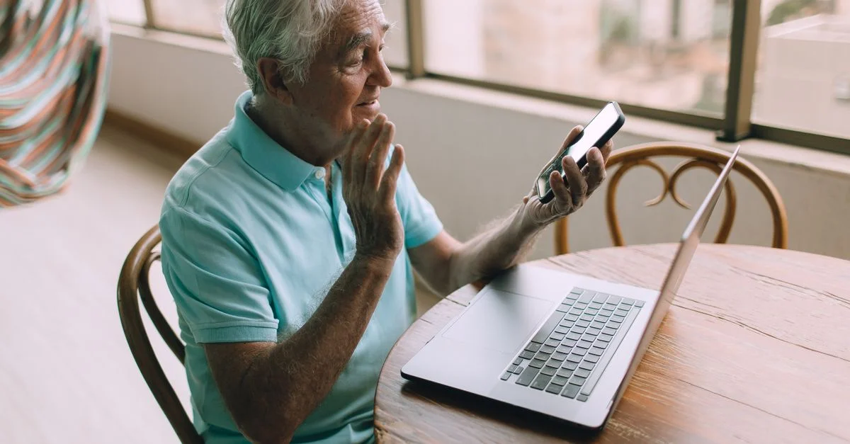 Un homme senior participant à un appel vidéo en utilisant un smartphone et un ordinateur portable à l'intérieur.