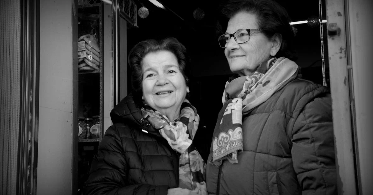 Portrait en noir et blanc de deux femmes âgées souriant devant un magasin.